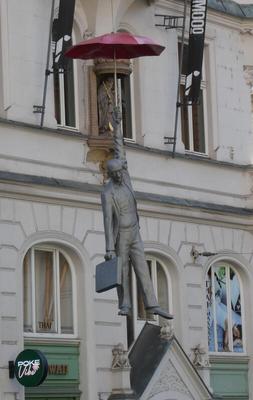 Statue of man with briefcase in one hand. He is hanging from a red  umbrella in the other hand.
